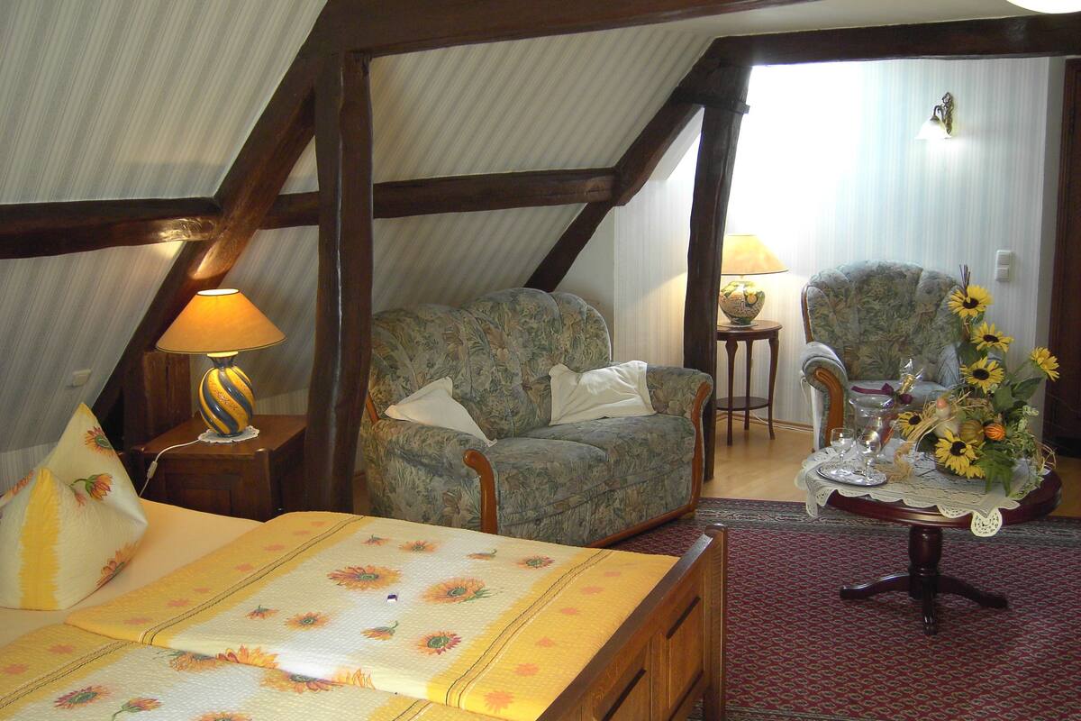 A bedroom with a bed, a floral armchair, a small table with a lamp, and a vase with sunflowers.