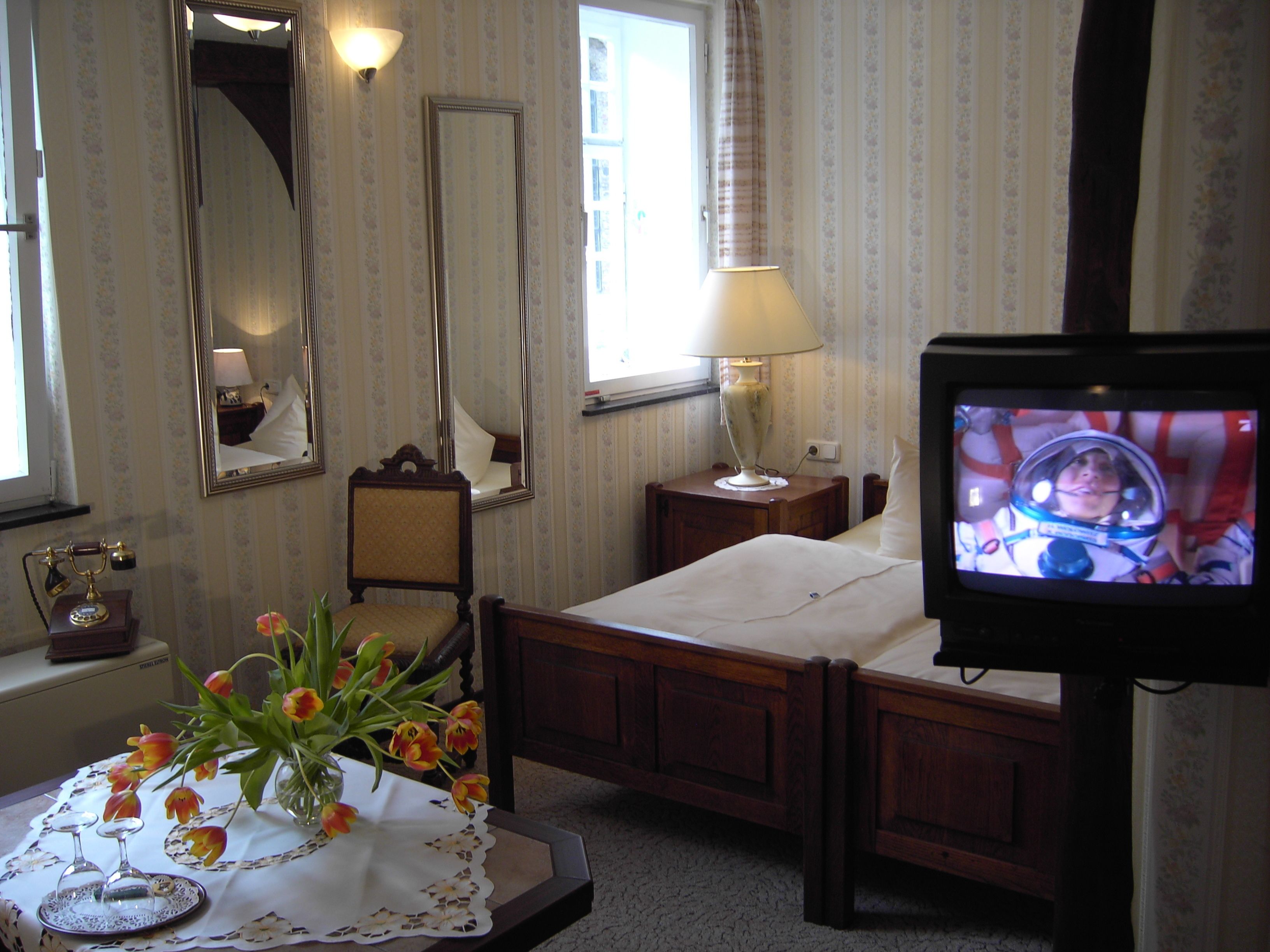 Ein Hotelzimmer mit Bett, Fernseher, Nachttisch mit Lampe, Sessel, Spiegel und Blumen in einer Vase.