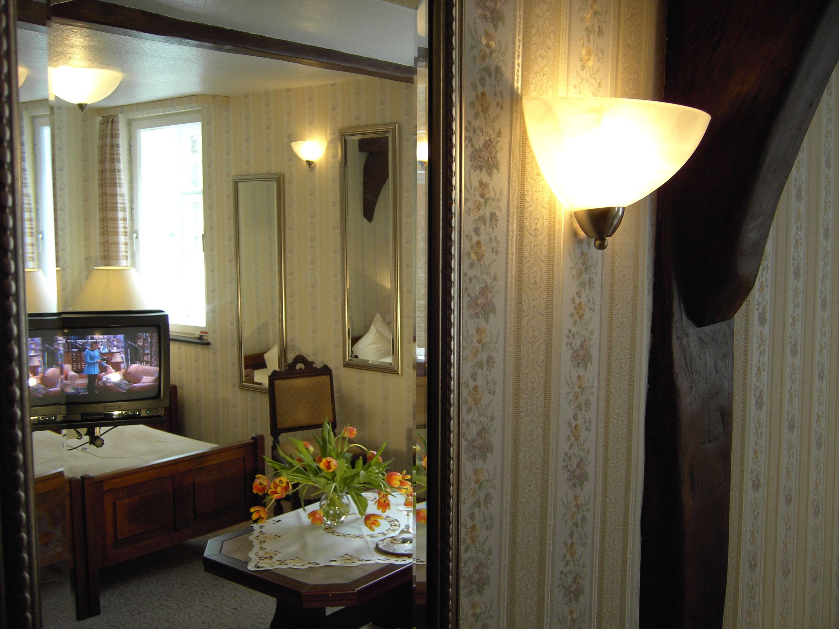 Une chambre avec un lit, une table de chevet ornée d’un vase de fleurs, une télévision et un miroir.
