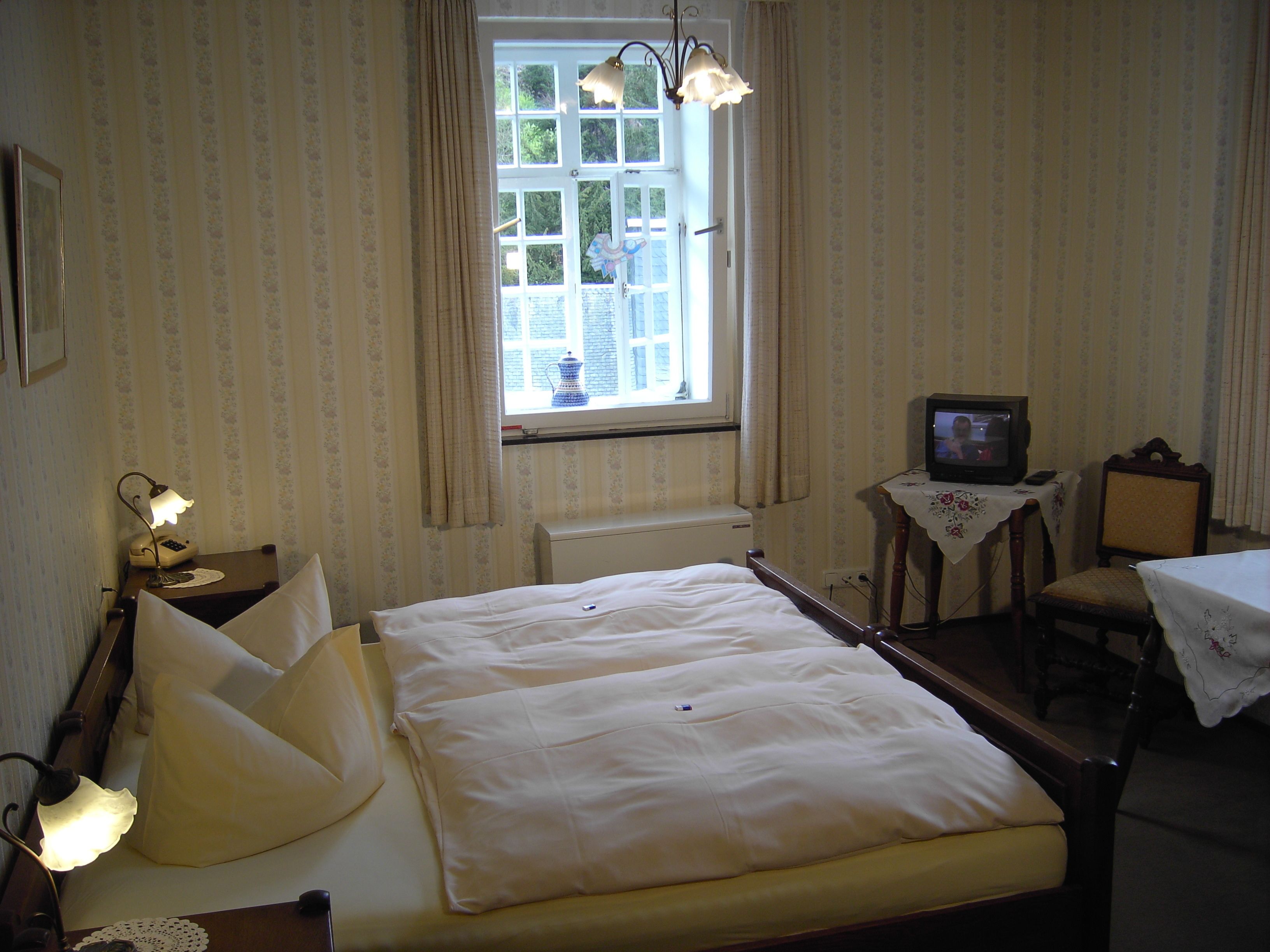 A hotel room with a bed, a television, a small table with a floral tablecloth, and a window with curtains.