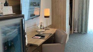 A hotel room with a desk, a chair, a television, a lamp, and a minibar. - Stadthotel Geis (Bad Neustadt an der Saale)