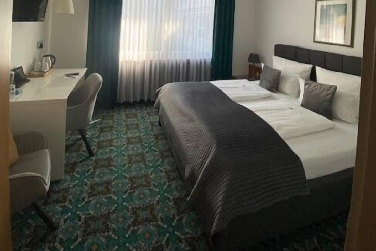 A hotel room with a bed, a desk with a chair, a television, and a window with curtains.