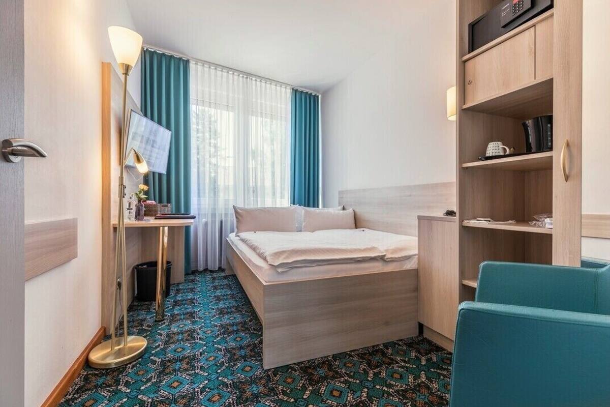 A compact hotel room with a bed, a desk, a chair, a TV, and a shelf with a microwave.