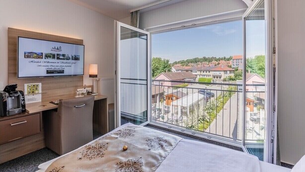 A hotel room with a bed, a desk with a coffee maker, a TV on the wall, and a large window with a view of buildings and trees. - Stadthotel Geis (Bad Neustadt an der Saale)