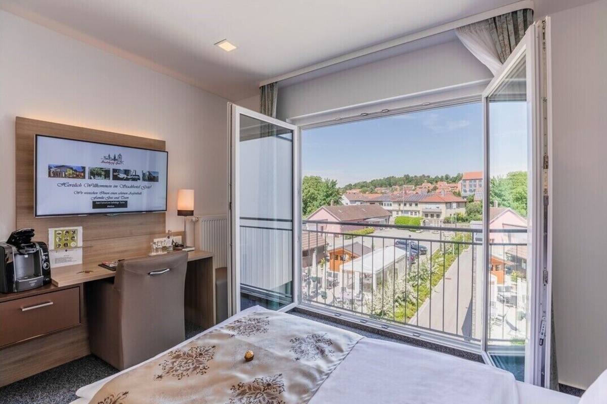 A hotel room with a bed, a desk with a coffee maker, a TV on the wall, and a large window with a view of buildings and trees.