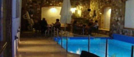 Zona de piscina exterior con paredes de piedra, algunas personas sentadas relajándose y un área de estar cubierta.