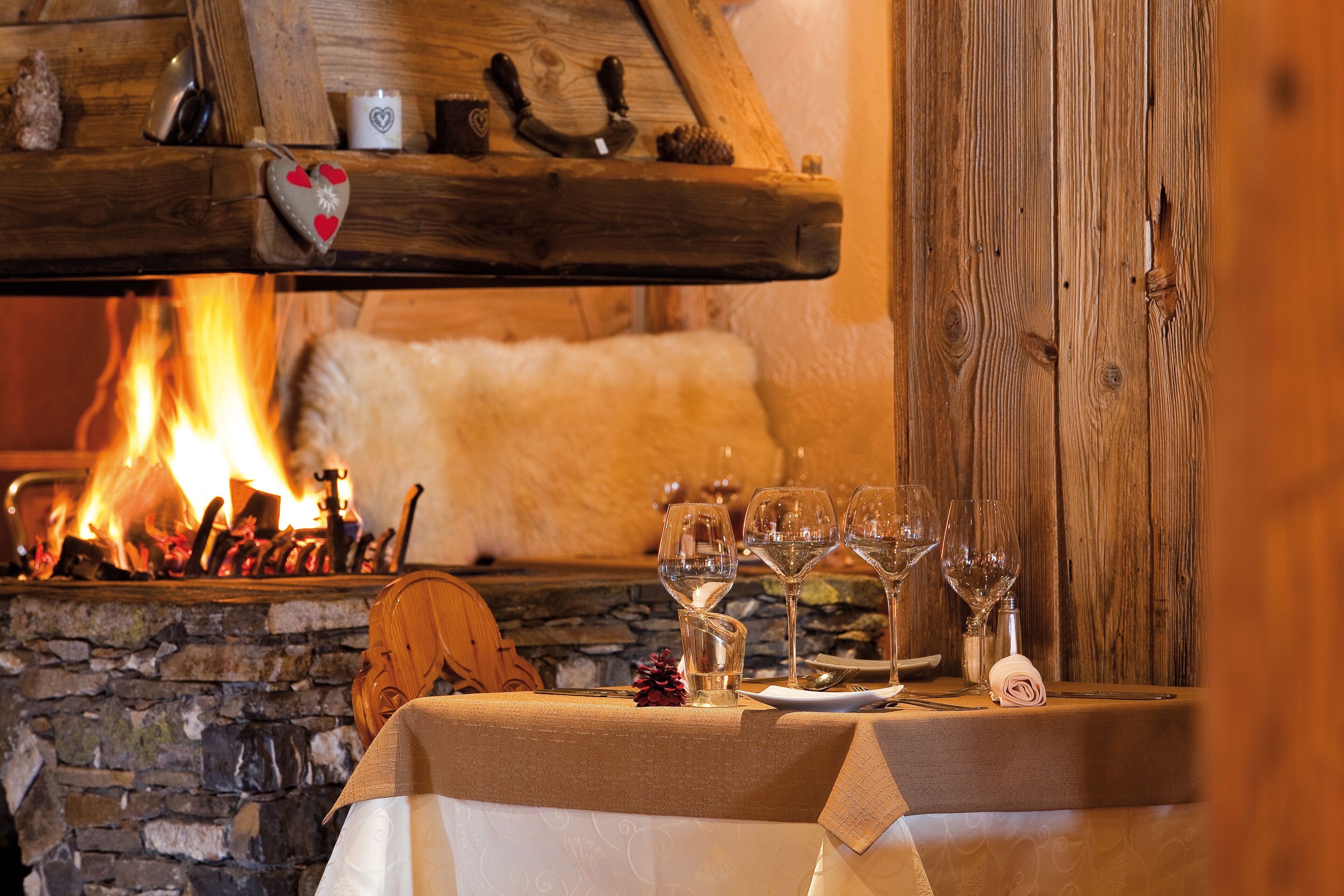 A table set for dining with wine glasses and a candle, a fireplace with a stone surround, and wooden decor.