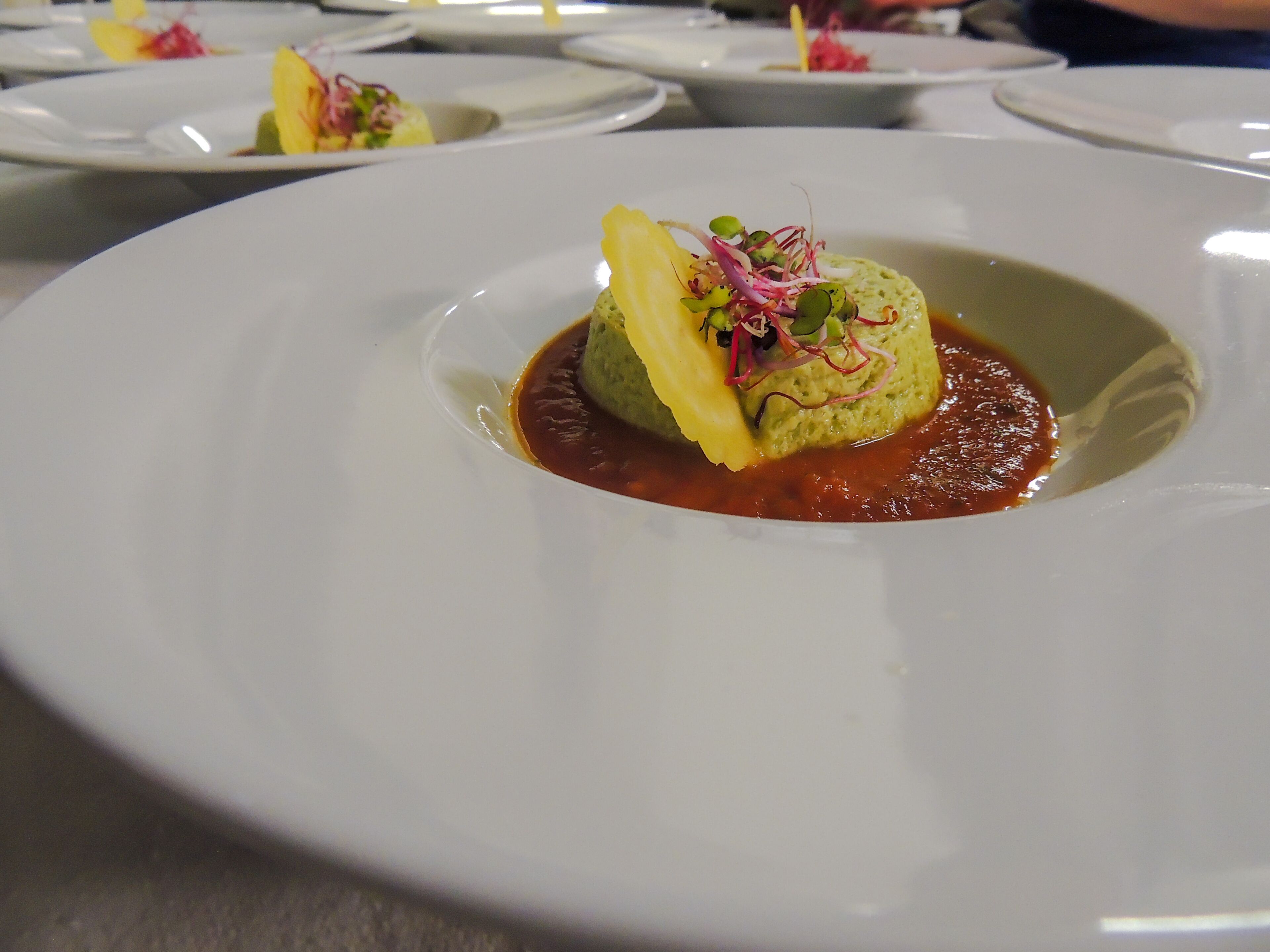 A white plate with a green mound of food topped with a slice of yellow vegetable and red vegetable garnish, surrounded by a red sauce.