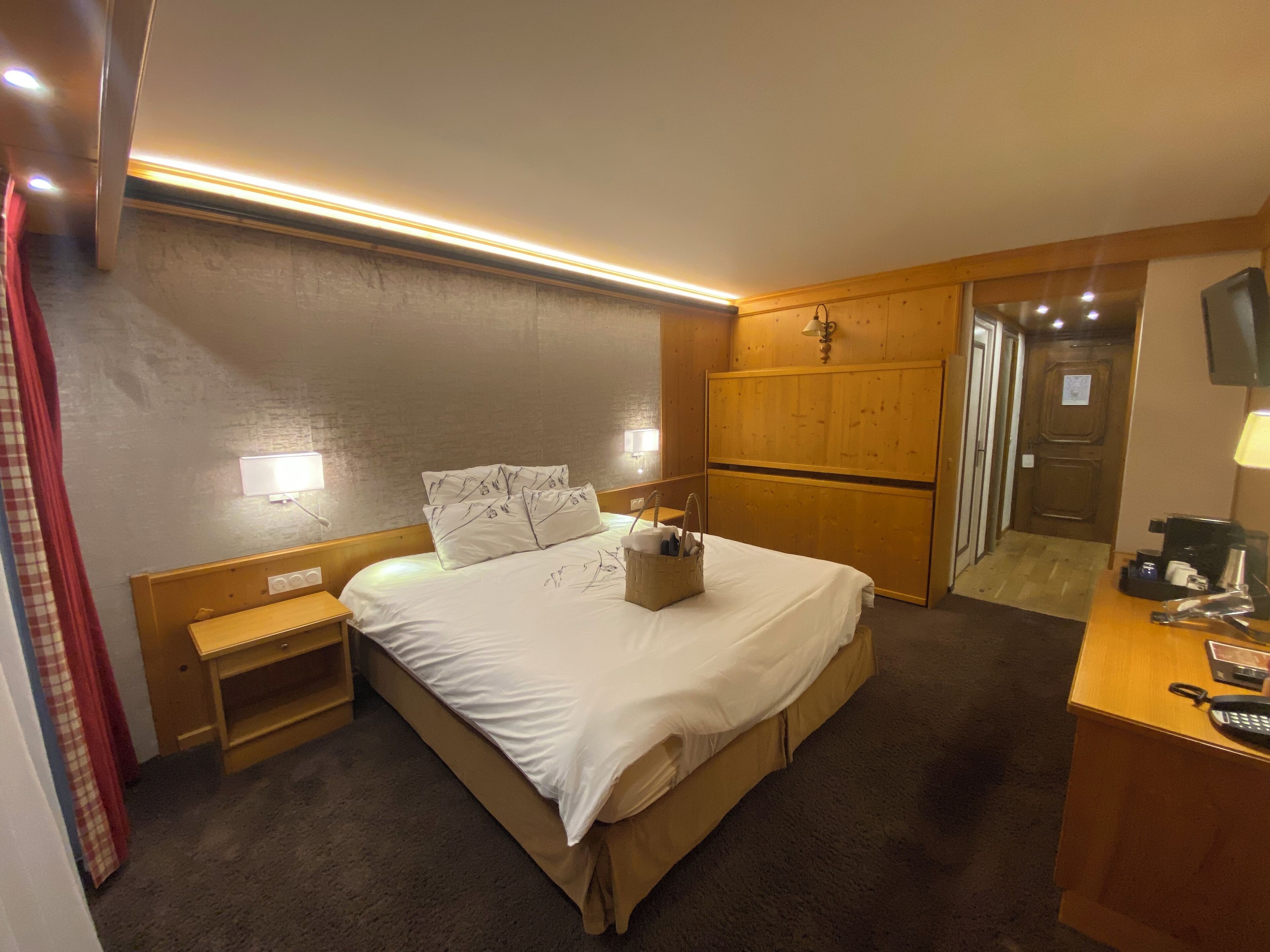 A hotel room with a bed, bedside tables, a wooden wardrobe, a desk with a telephone, and a television.