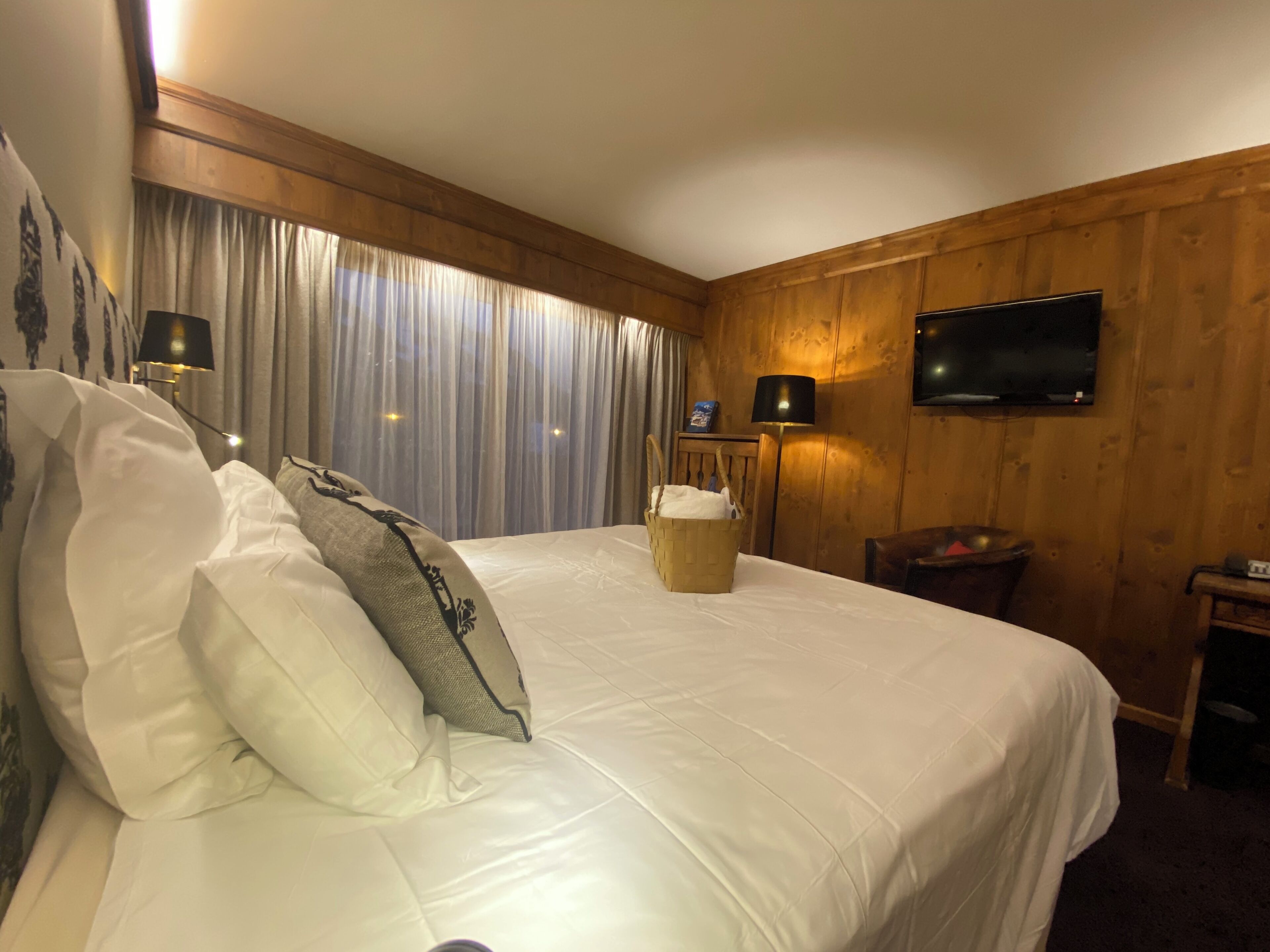A hotel room with a large bed, wooden paneling, a flat-screen TV, and a nightstand with a lamp.
