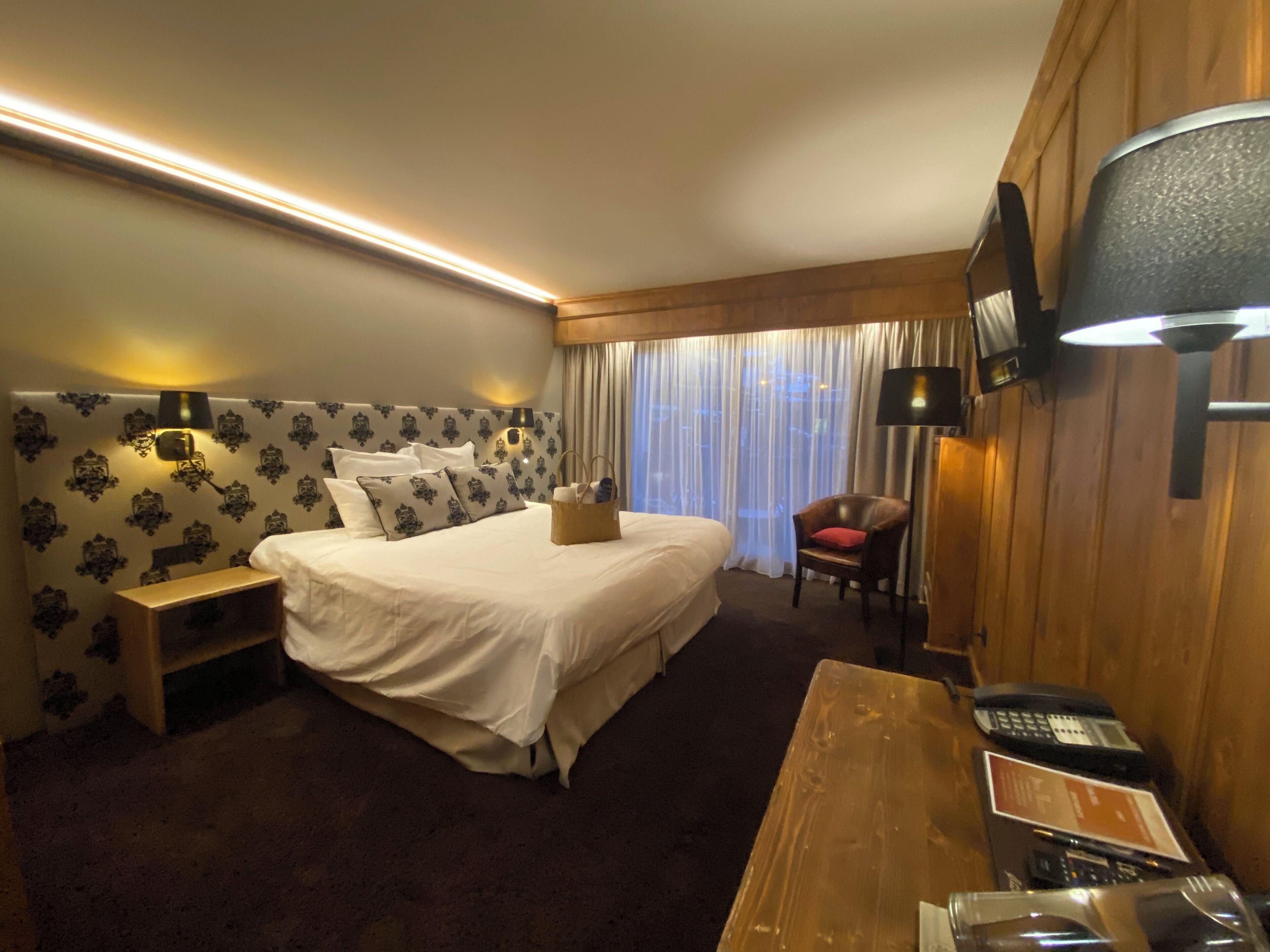Ein Hotelzimmer mit einem großen Bett, Nachttischen, einem Holztisch mit Telefon und einem an der Wand montierten Fernseher.