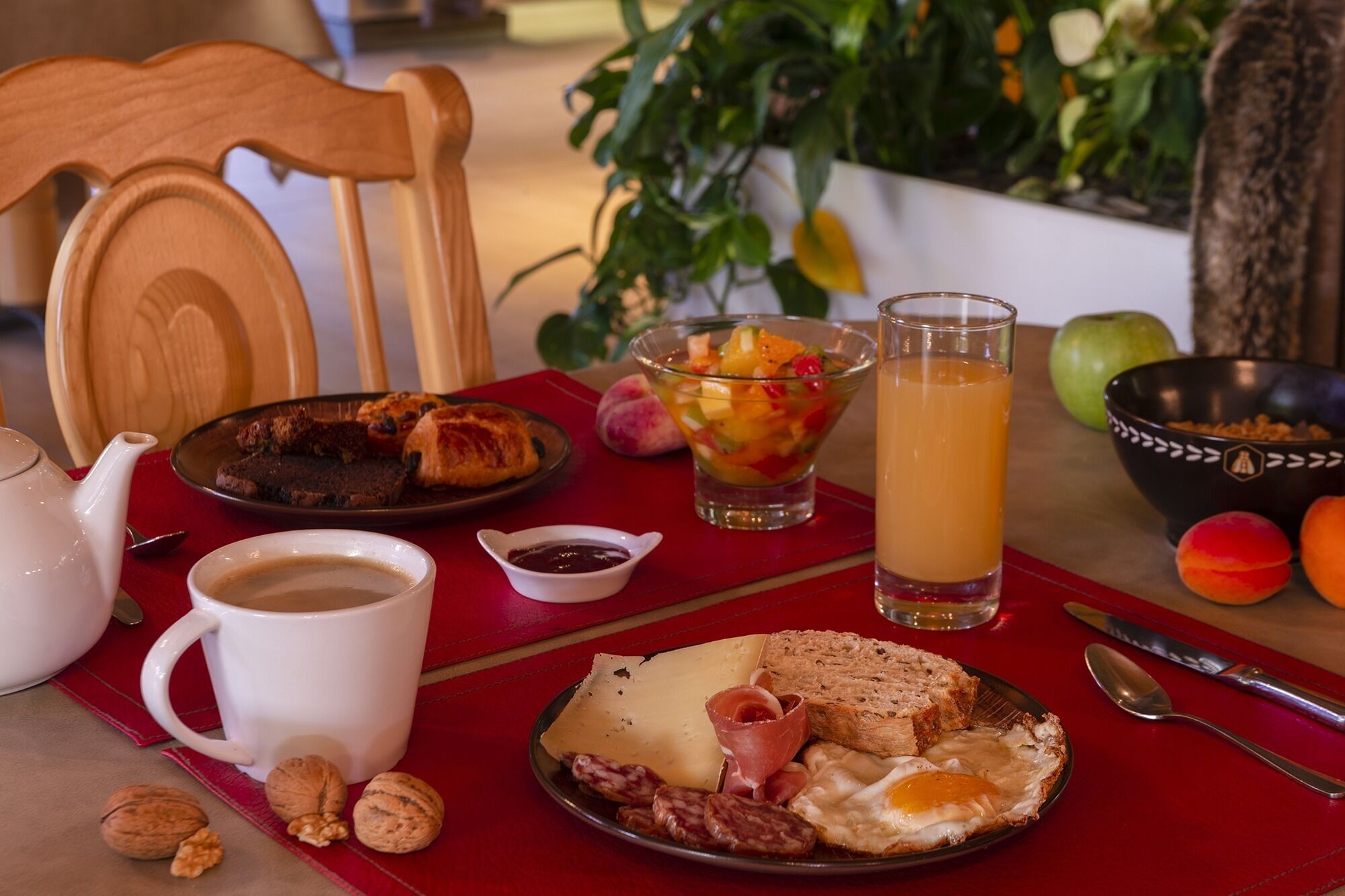A breakfast table with a plate of eggs, sausages, cheese, and bread, a glass of orange juice, a cup of coffee, and a bowl of fruit salad.