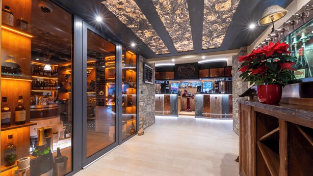 A bar area with a wooden counter, shelves with bottles, and a large potted plant. - Hôtel Macchi Restaurant & Spa (Chatel)
