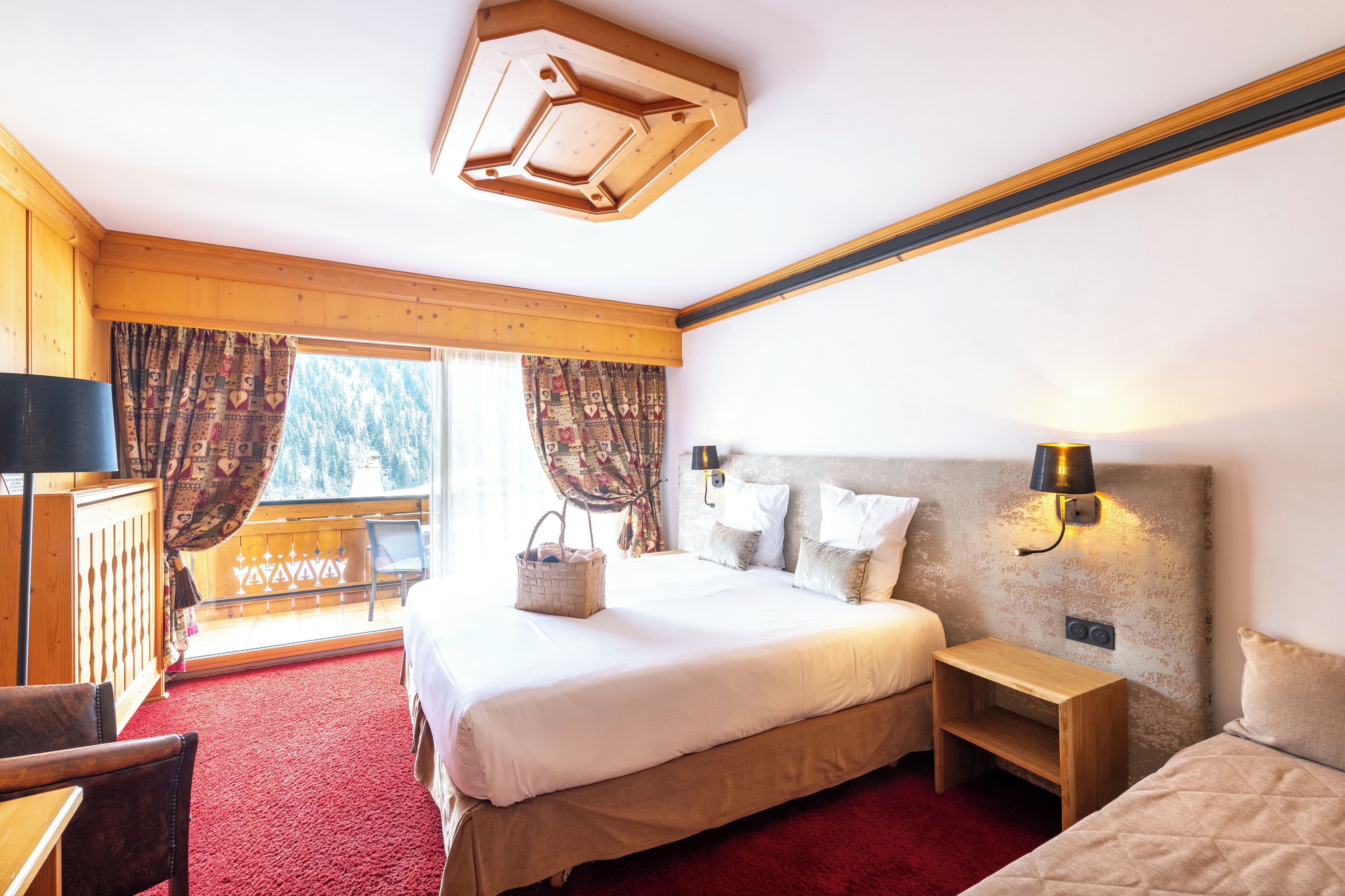 Une chambre d’hôtel avec un grand lit, une table de chevet en bois, un bureau et une chaise. Il y a une fenêtre avec des rideaux et une vue sur les arbres à l’extérieur.