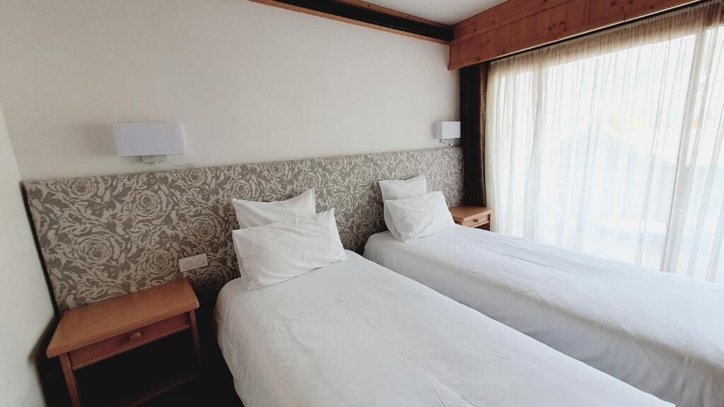 A hotel room with two single beds, a patterned headboard, bedside lamps, and a window with sheer curtains.