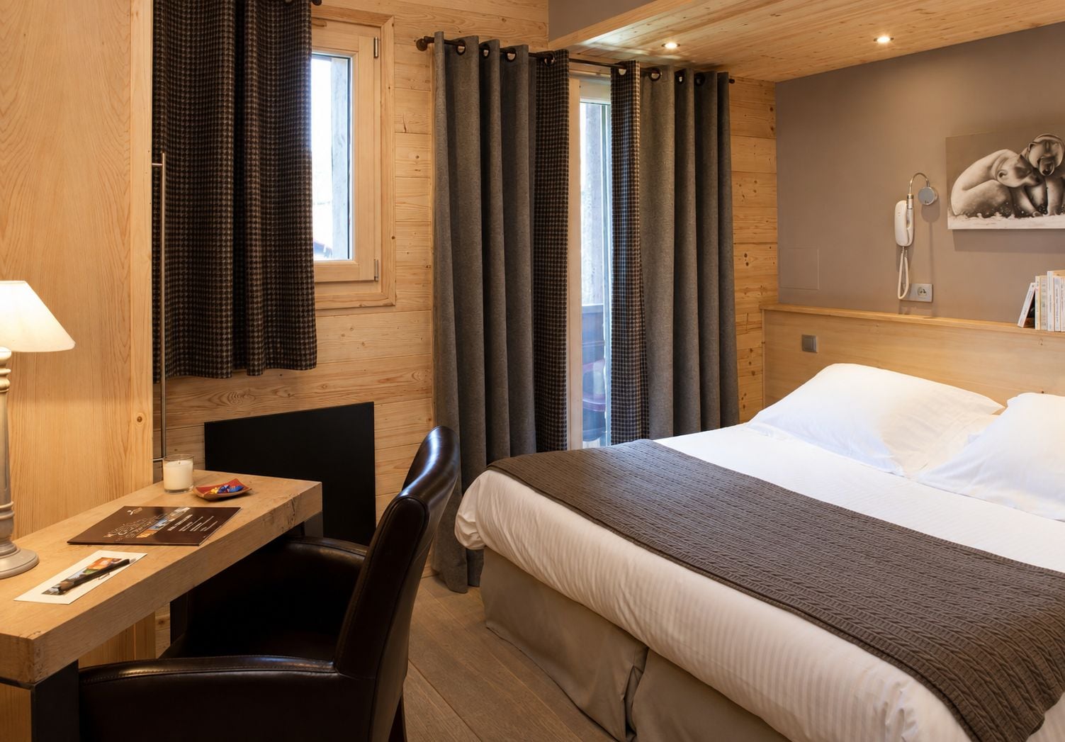 A hotel room with a bed, a desk with a chair, a window with curtains, and a wall-mounted phone.