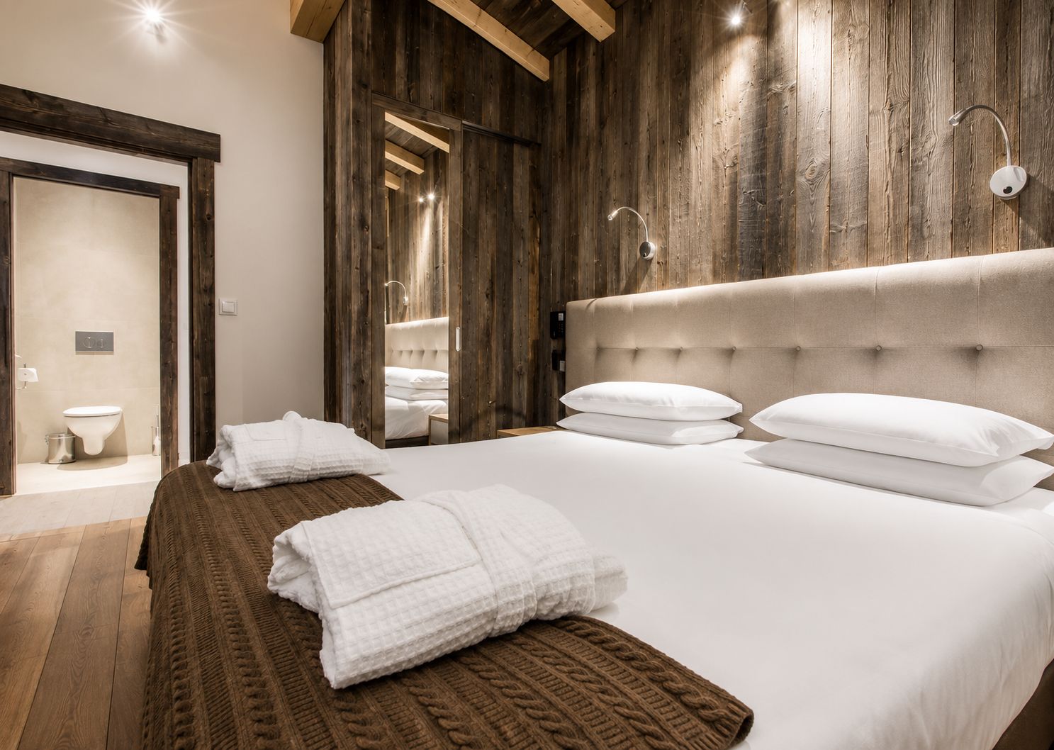 Une chambre d’hôtel avec un grand lit, des murs en bois, une tête de lit moderne et une salle de bain visible.