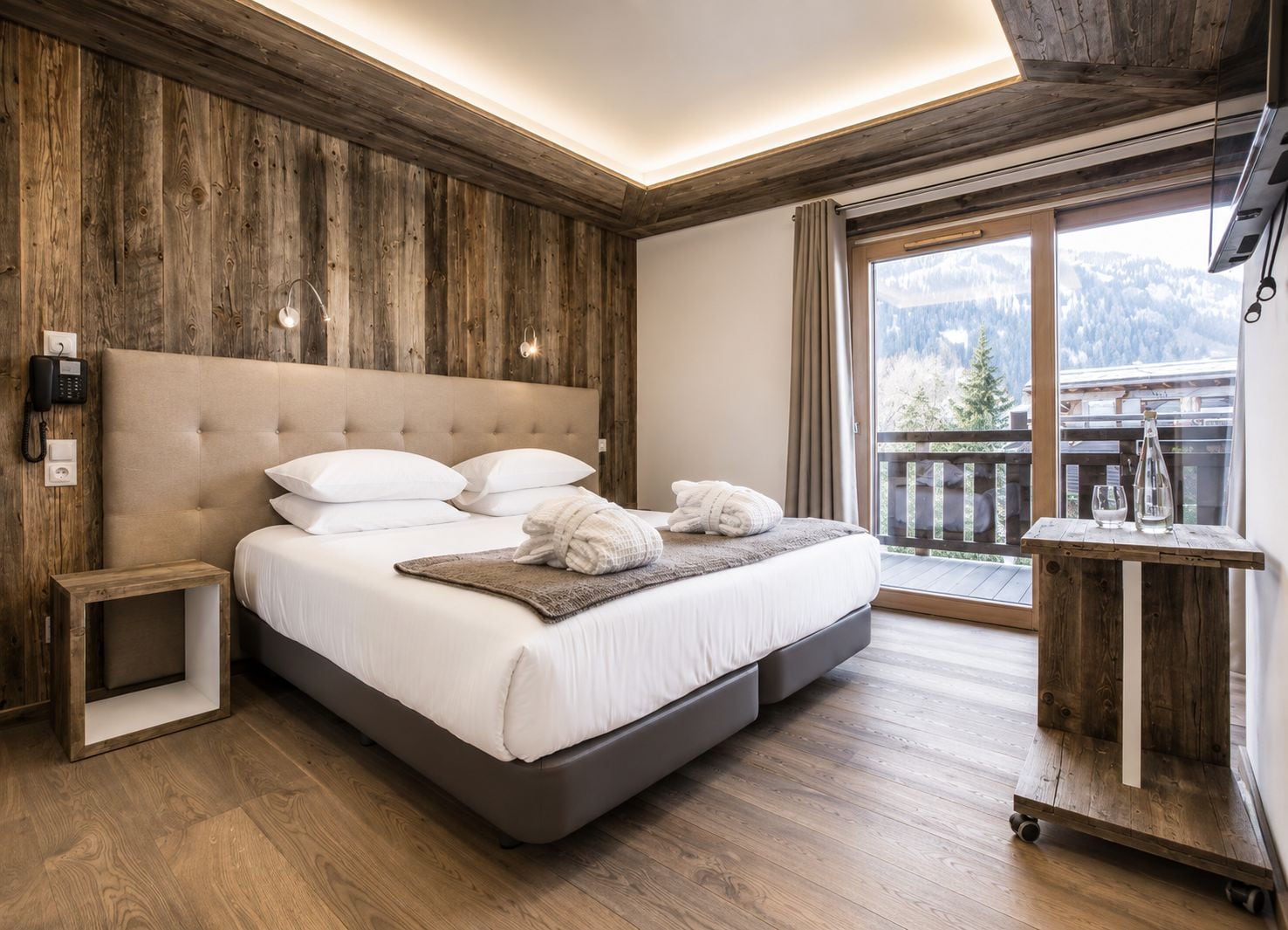 Una camera da letto con un letto grande, pareti e pavimento in legno, un comodino, un telefono e vista su un balcone con montagne sullo sfondo.