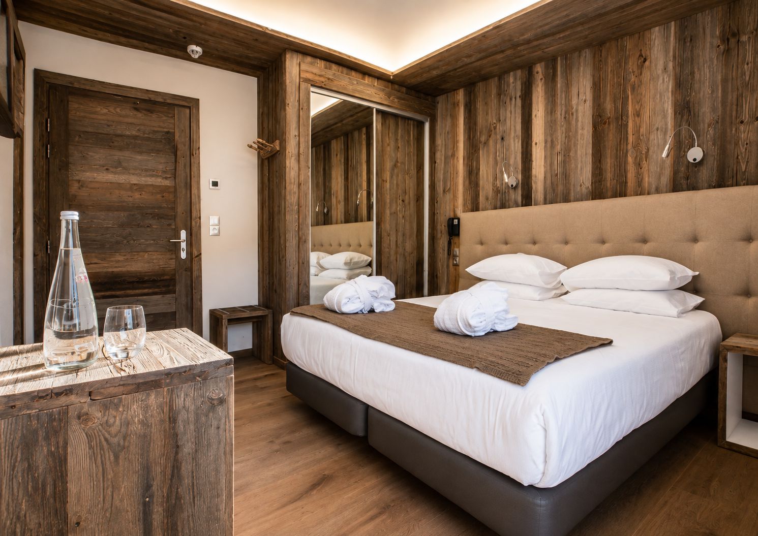 A hotel room with a wooden floor, a large bed with white linens, a bedside table with a lamp, and a wooden wardrobe.