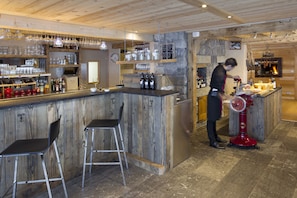 Un coin bar au style rustique, avec des murs en bois, un mur d’accent en pierre et un comptoir. On y trouve des tabourets, un présentoir à bouteilles de vin et une personne qui travaille derrière le bar.