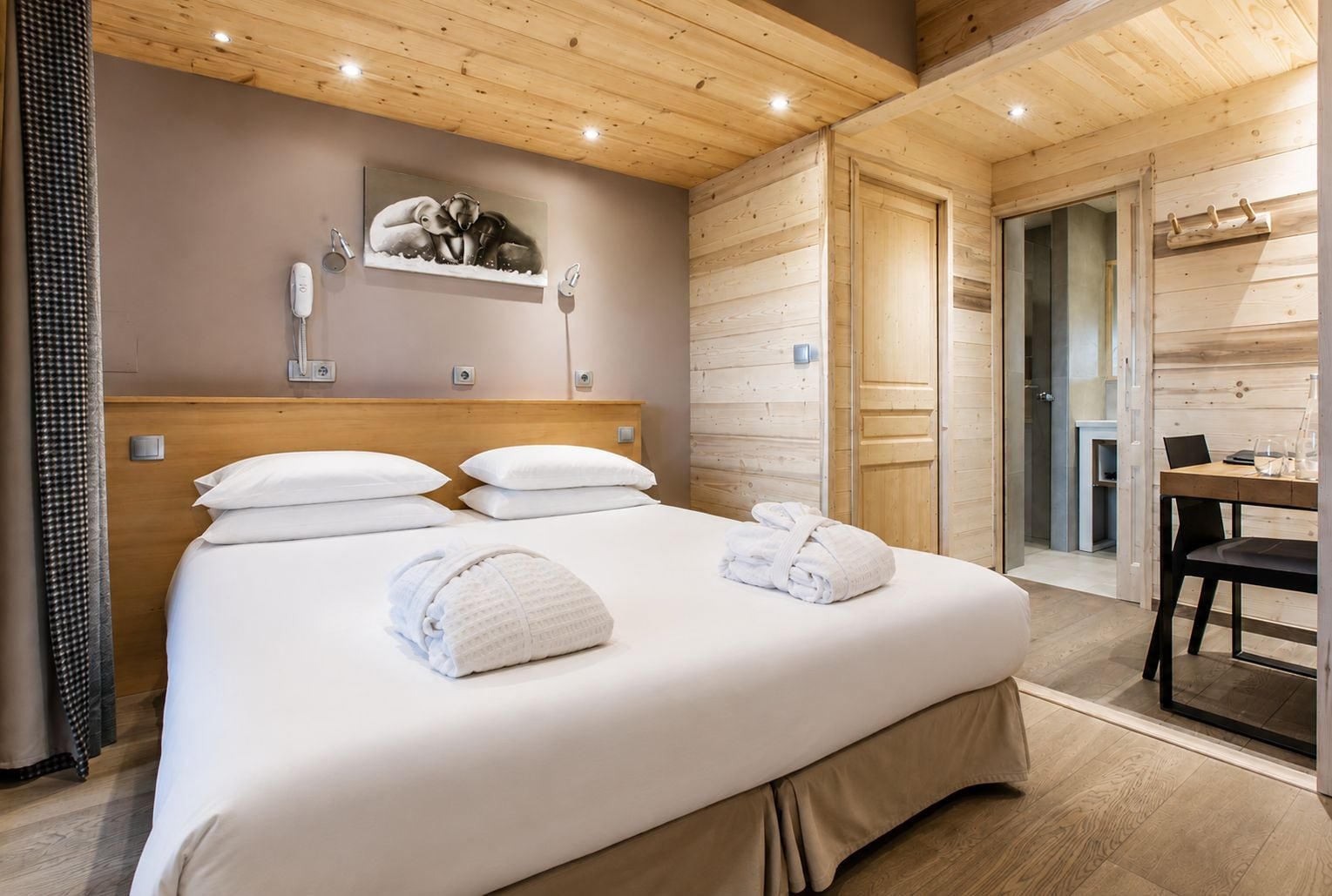 A hotel room with a wooden ceiling, a bed with white linens, a painting on the wall, and a bathroom visible through an open door.