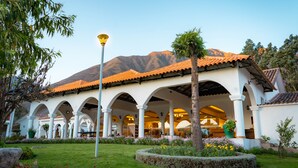 A building with white arches and a tiled roof, surrounded by a well-maintained garden with flowers and a tree. - Hotel Agustos Urubamba (Urubamba)