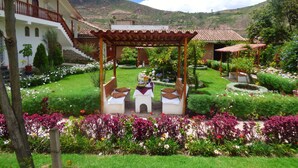 A garden with a gazebo, a white building with a red roof, and a mountainous backdrop. - Hotel Agustos Urubamba (Urubamba)