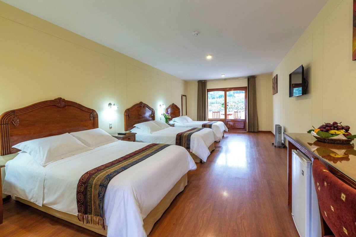 A hotel room with three beds, wooden headboards, bedside lamps, a flat-screen TV, a small refrigerator, and a window with a view of the outdoors.