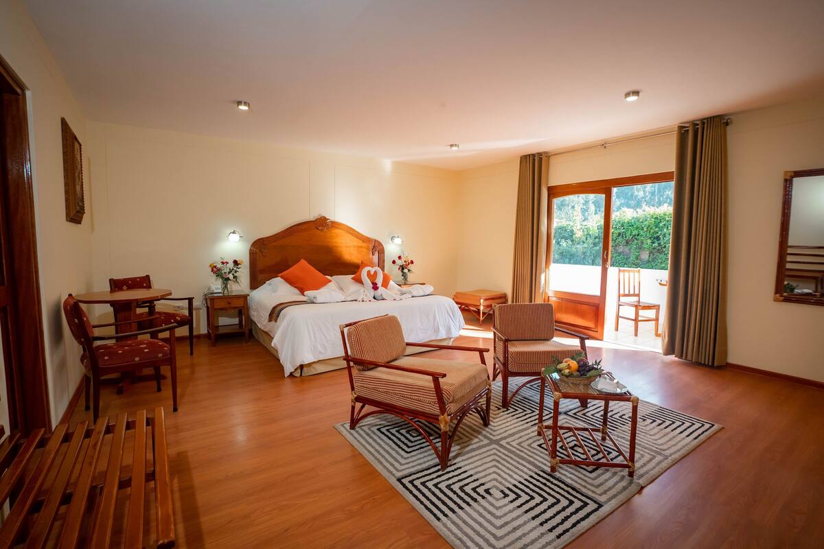 A hotel room with a large bed, wooden furniture, a dining table, and a balcony with curtains.