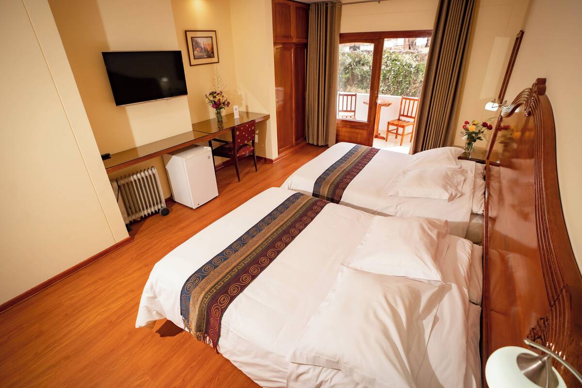 A hotel room with two beds, a desk, a chair, a TV, and a view of the outside.