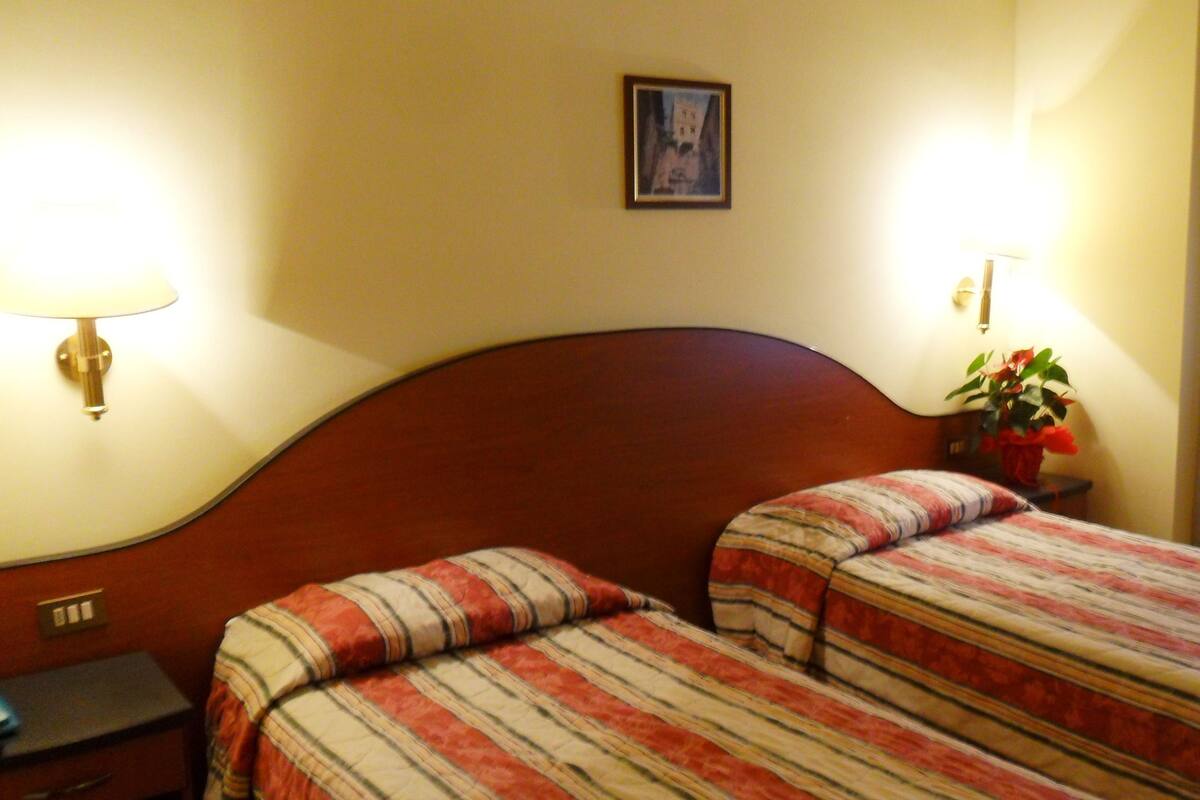 A hotel room with two single beds, a wooden headboard, a nightstand with a lamp, a framed picture on the wall, and a potted plant.