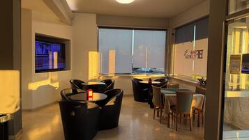 A modern hotel room with a large window, a flat-screen TV mounted on the wall, and a seating area with black chairs and a small table.