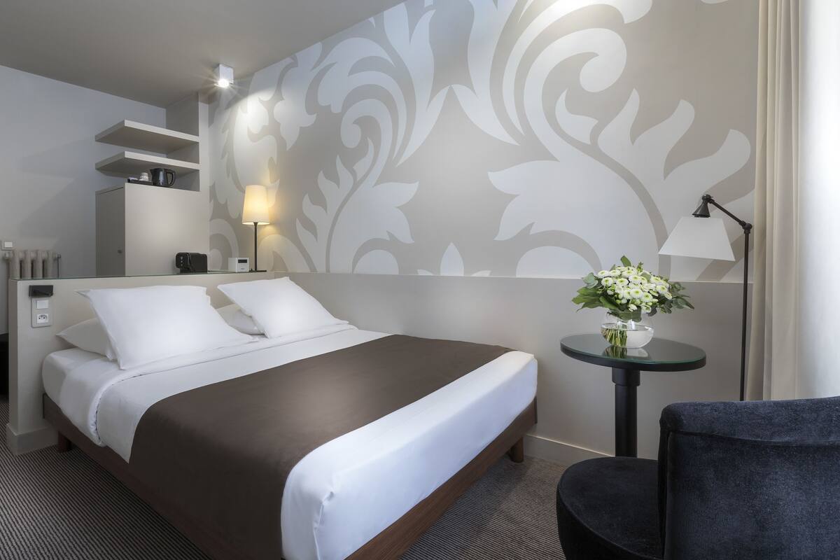 Quarto de hotel com cama, mesa de cabeceira, abajur e um detalhe decorativo na parede.