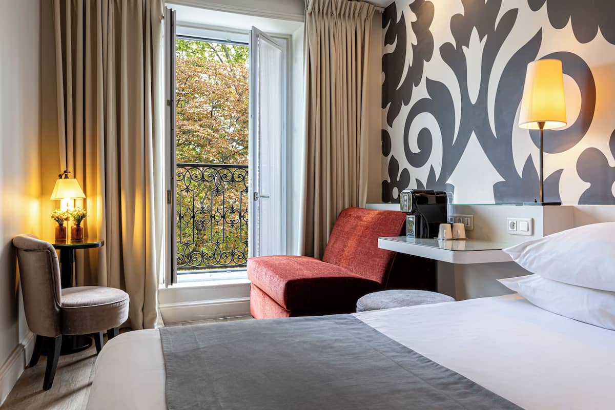 Quarto de hotel com cama, escrivaninha com luminária, cadeira e vista para o exterior.