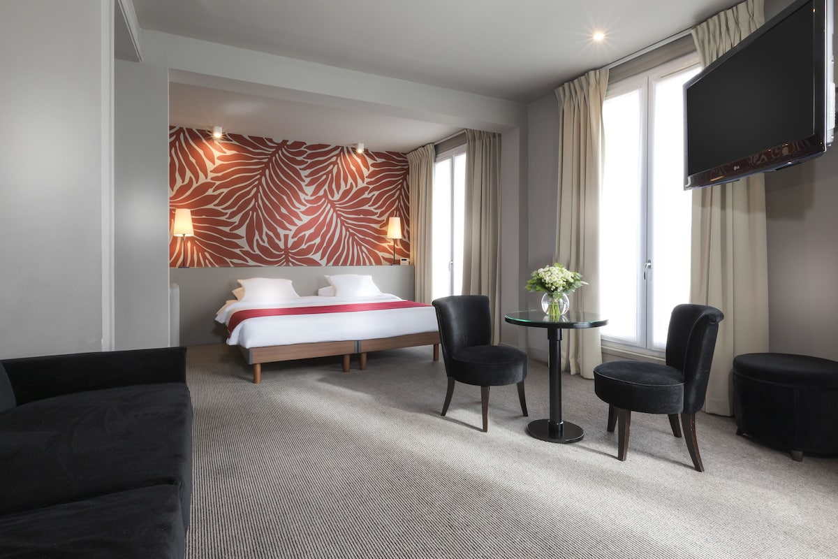 Quarto de hotel com uma cama, duas cadeiras, uma mesinha com um vaso, uma televisão e uma parede com padrão de zebra.