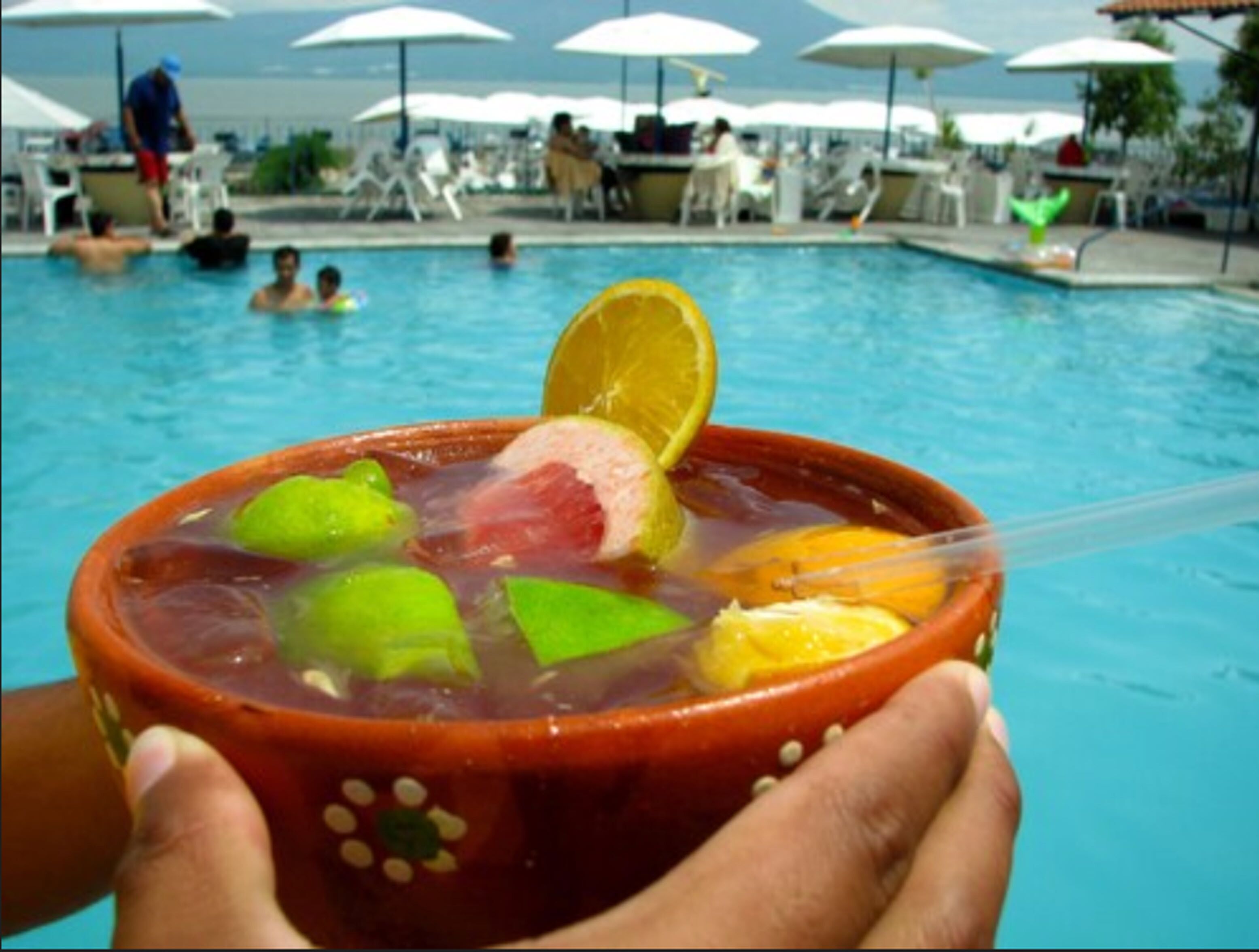 A person holding a bowl with a drink containing lime and fruit, in front of a pool area with umbrellas and people.