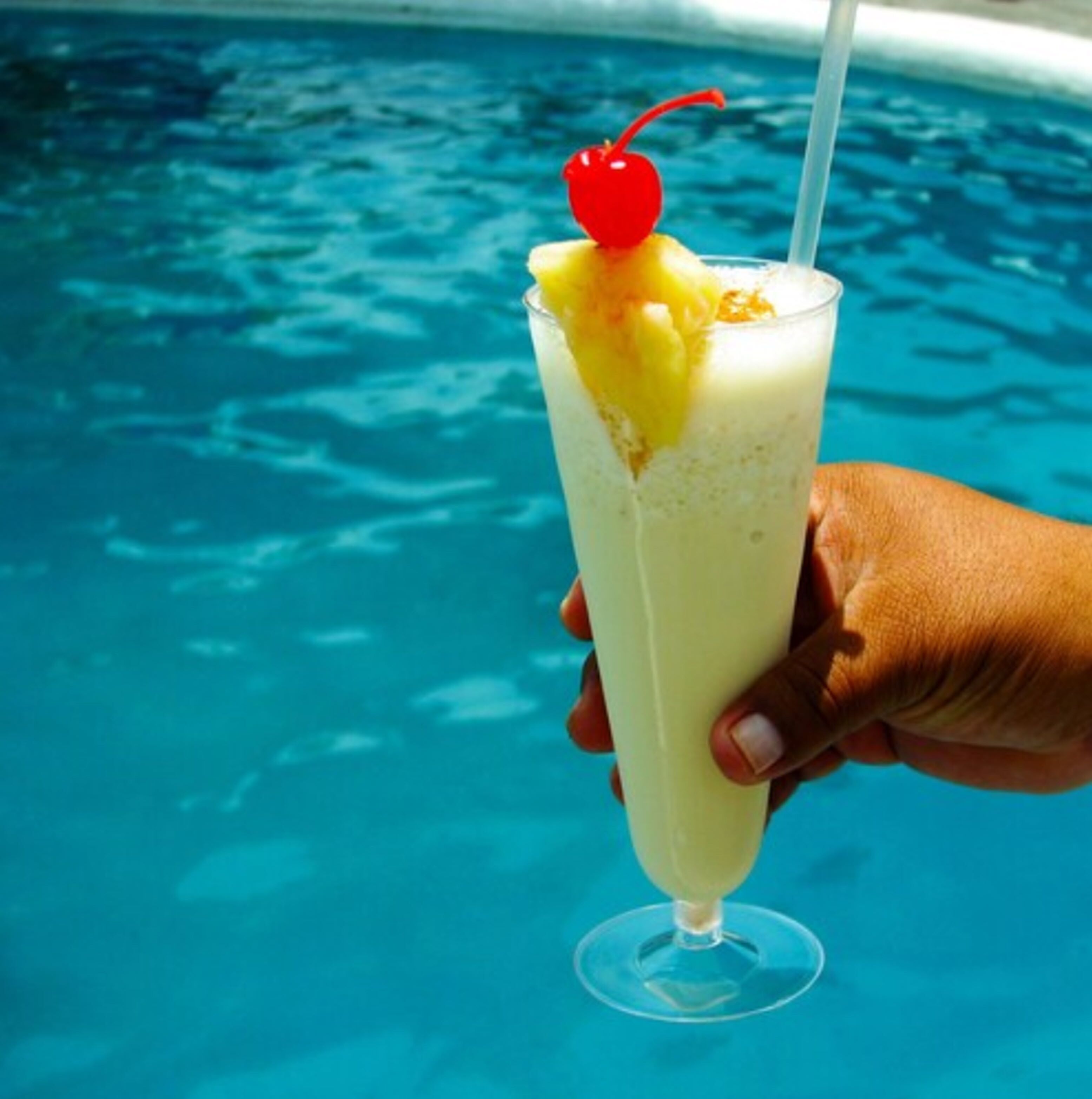 Una mano che tiene un bicchiere alto con una bevanda gialla, una ciliegina sopra e una cannuccia, con vista su una piscina.