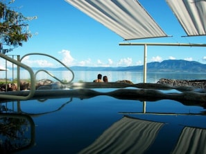 Un espace de baignade extérieur paisible, avec une eau d’un bleu limpide, une vue sur la mer et des montagnes à l’horizon.