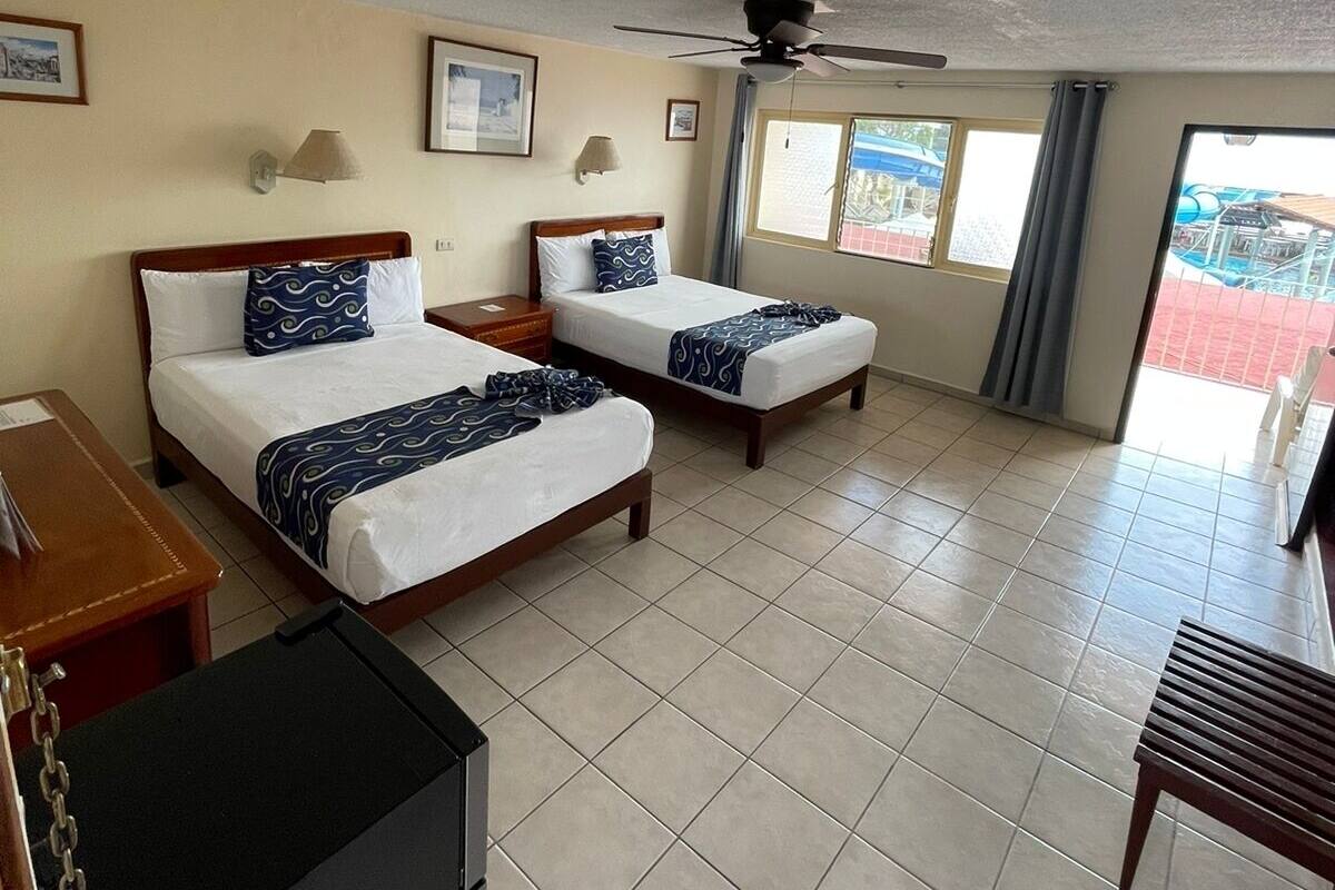 Ein Hotelzimmer mit zwei Betten, einem Schreibtisch, einem Stuhl, einem Deckenventilator und Blick auf einen Außenbereich.