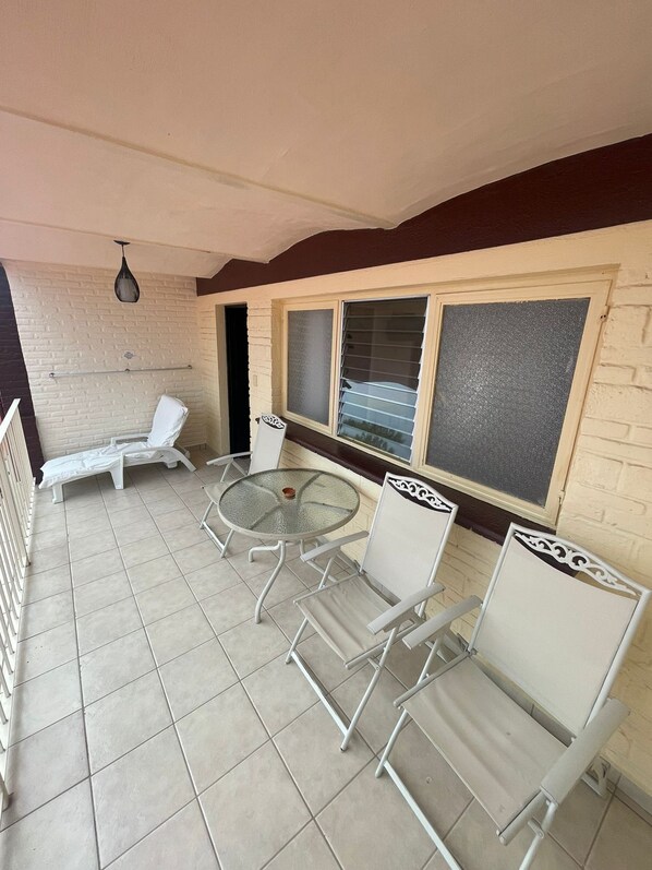 A covered patio area with a round glass table, white metal chairs, and two white lounge chairs.