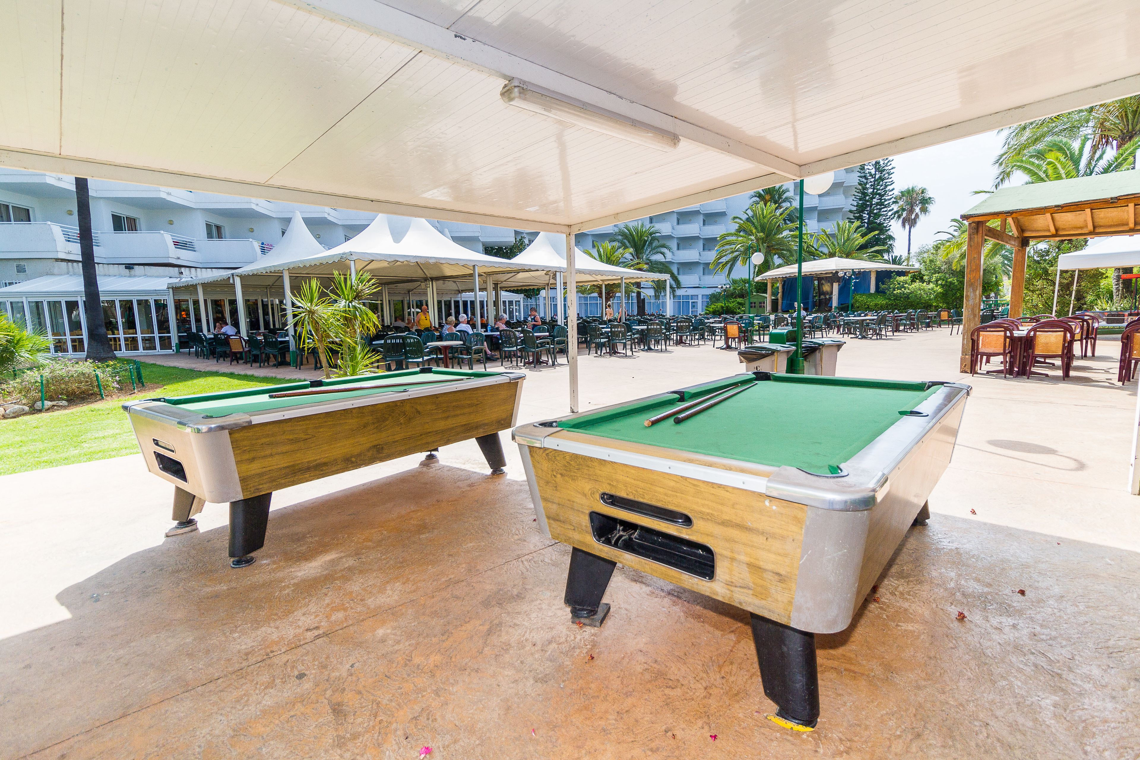 An outdoor pool area with two pool tables, covered seating, and a view of a hotel building in the background.