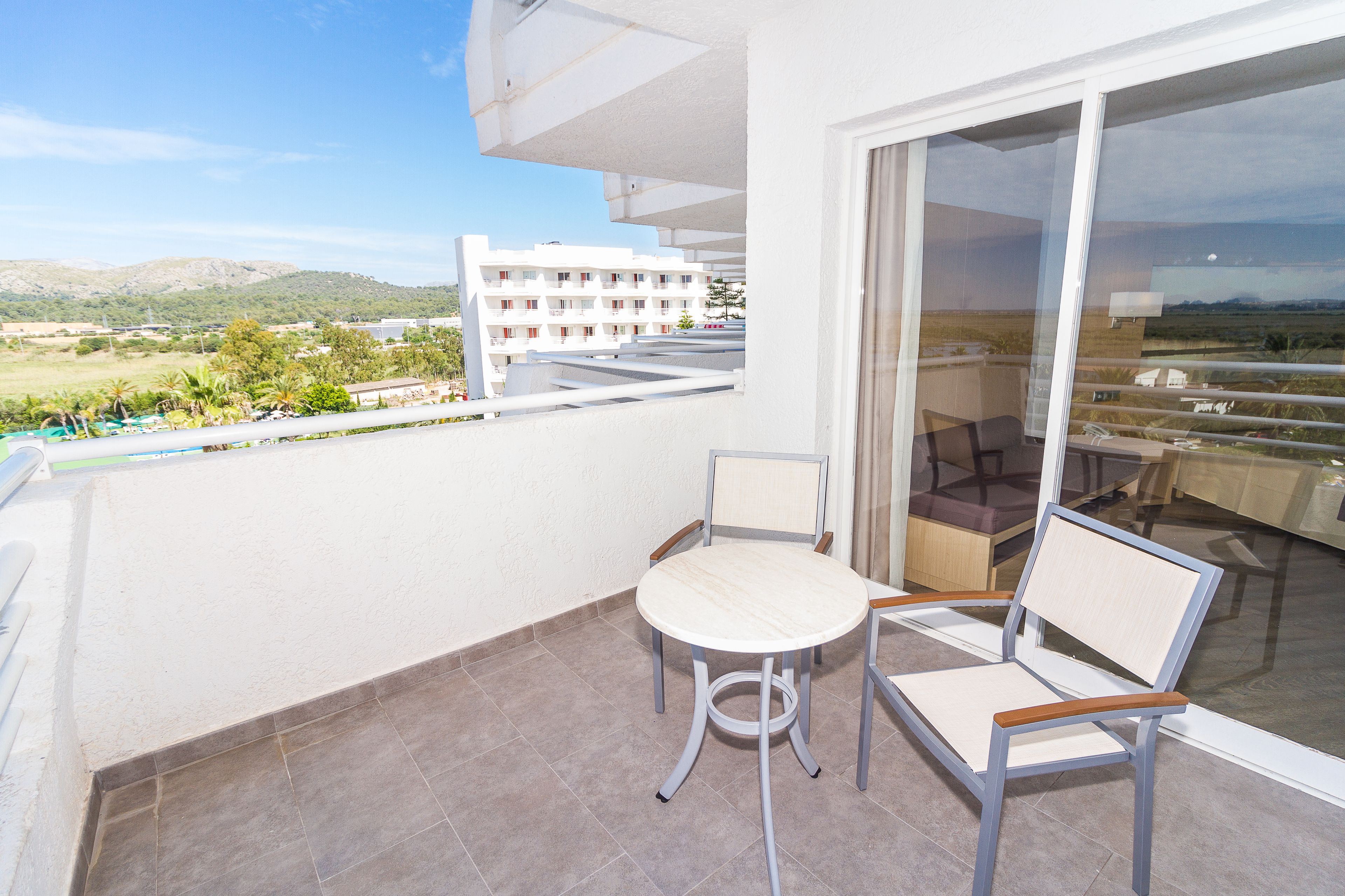 Un balcón con una mesa y dos sillas, con vistas a un edificio y un paisaje con árboles y colinas.
