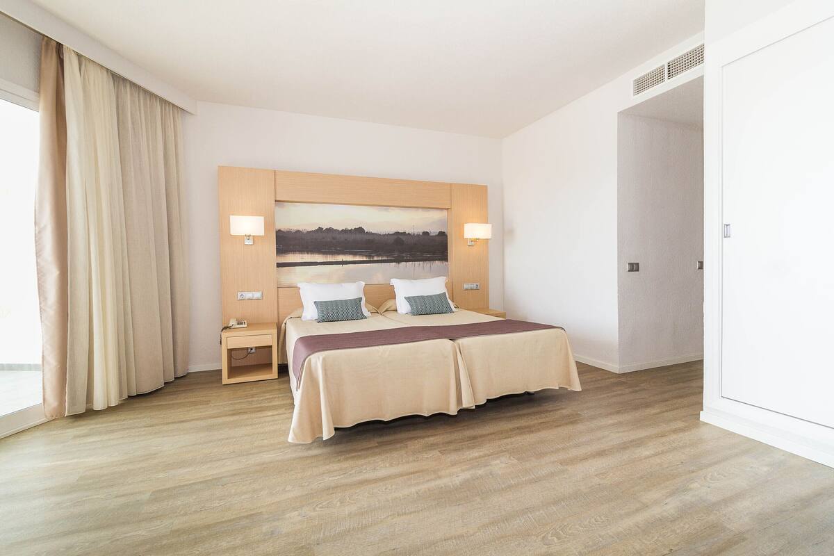 A hotel room with a large bed, a nightstand, a lamp, and a view of a lake through a window.