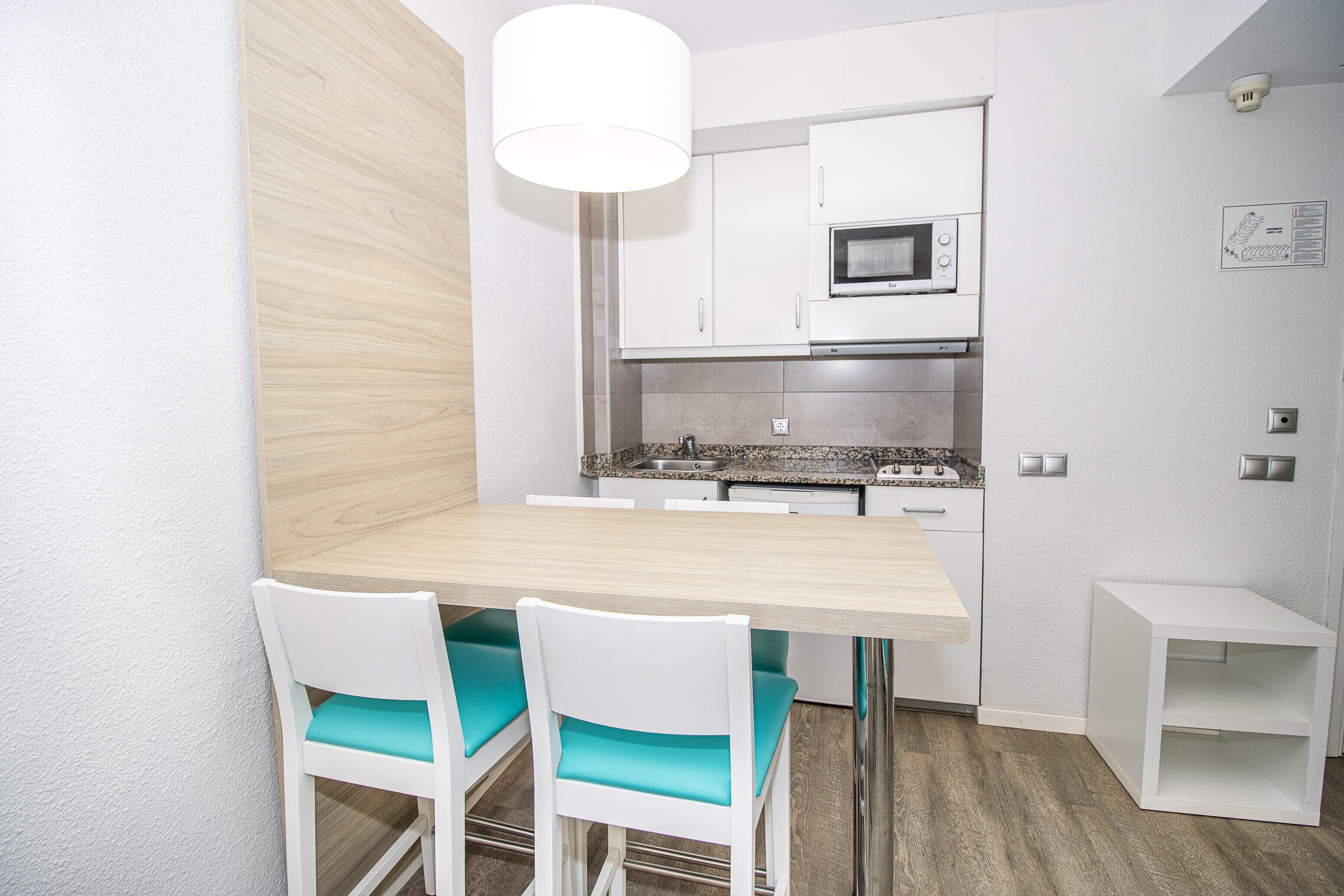 A modern kitchen with white cabinets, a microwave, and a stainless steel range hood. There are two white chairs with teal cushions and a white table.