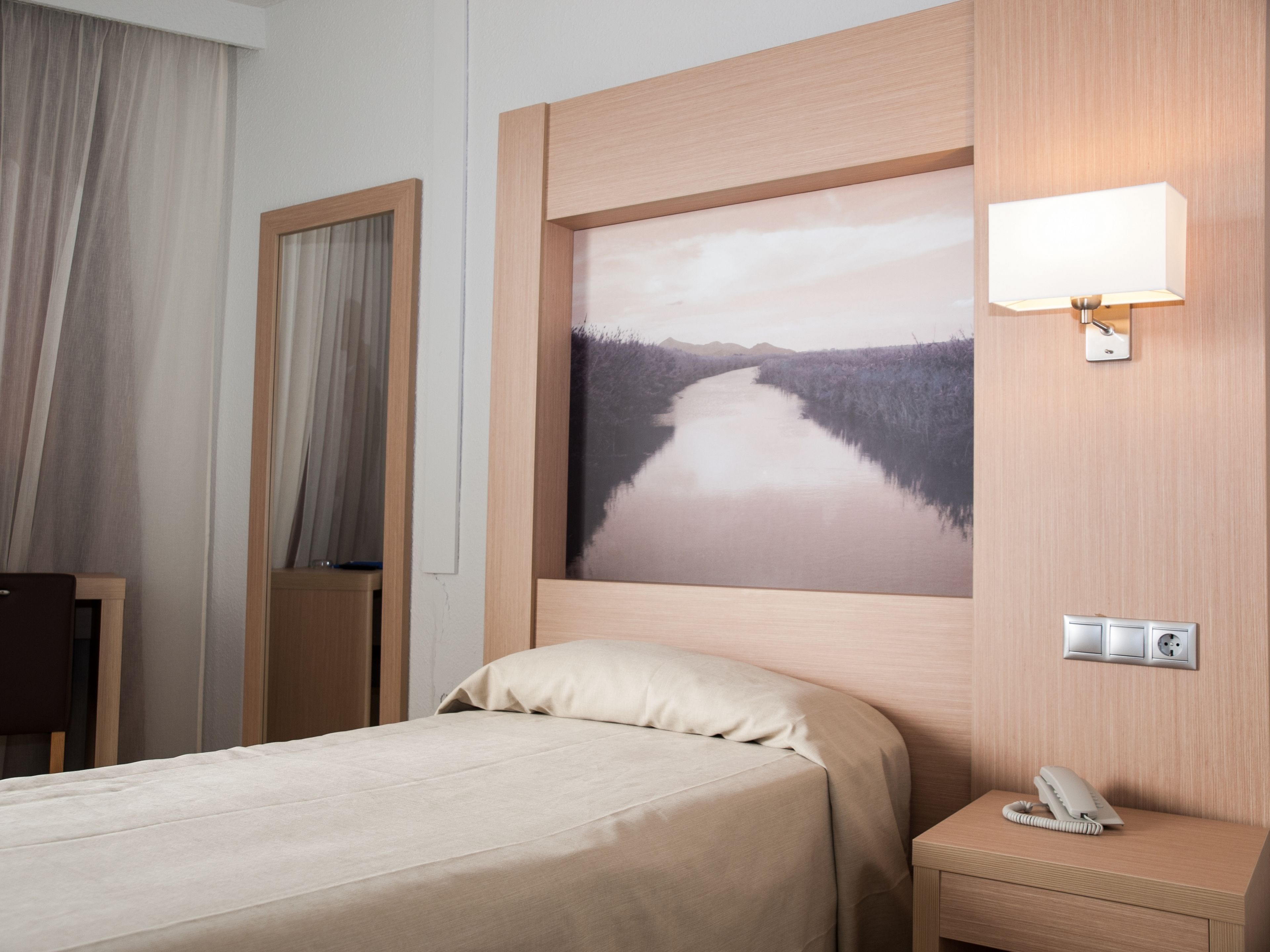 Ett hotellrum med en säng, ett nattduksbord med en telefon, en väggmonterad lampa och en stor, inramad tavla som föreställer en flod.