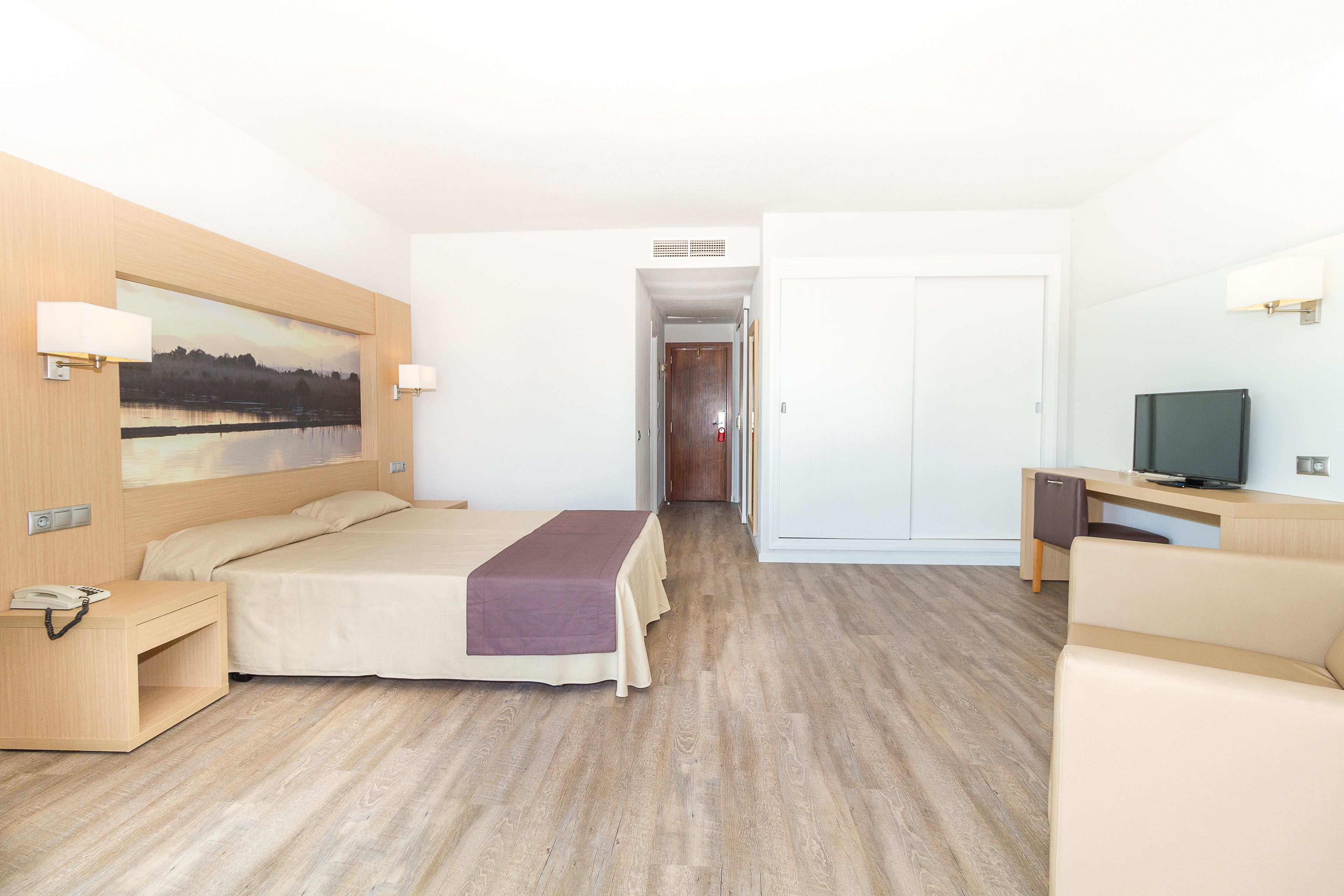 A hotel room with a bed, bedside table, lamp, TV, and a painting on the wall.