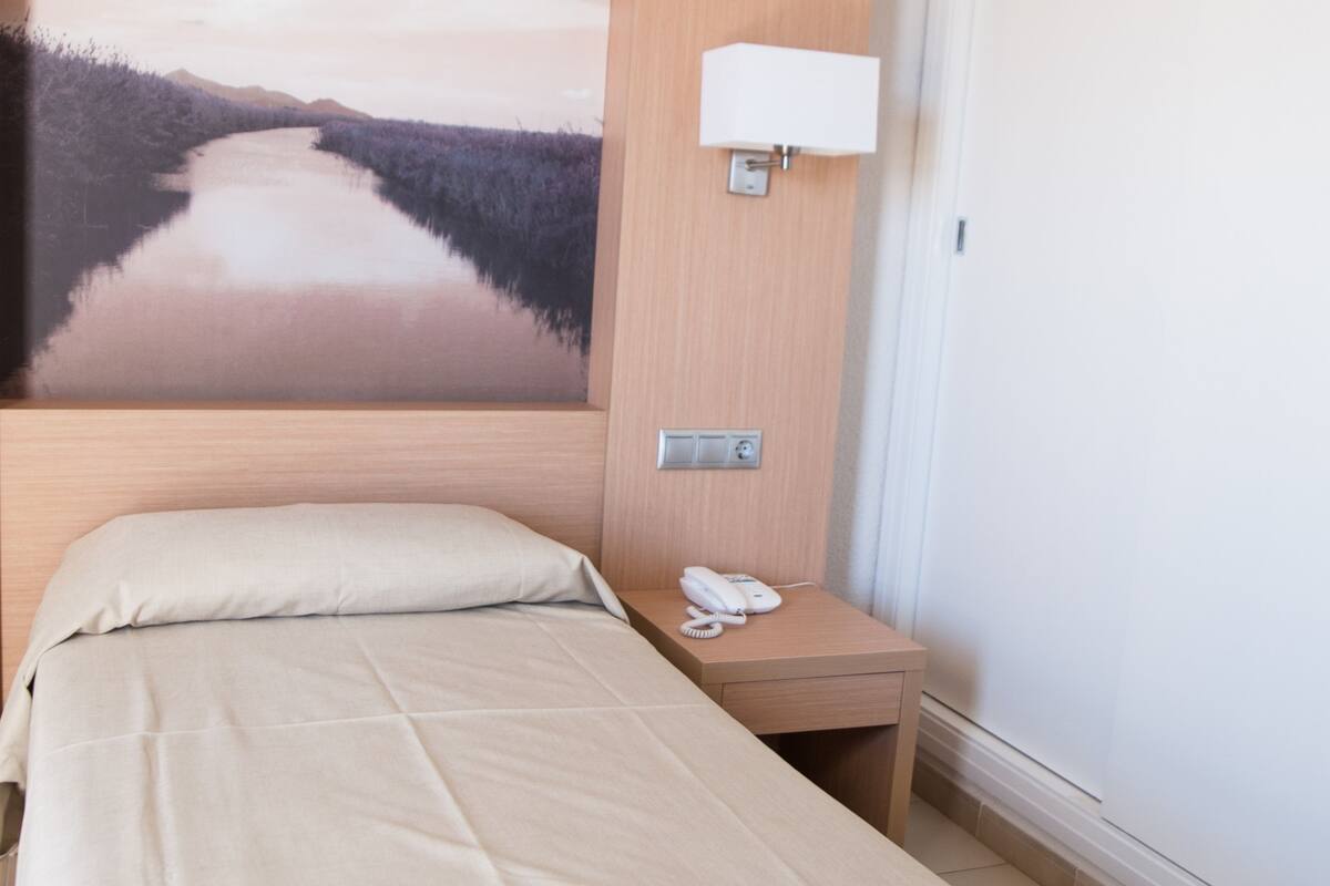 A hotel room with a bed, a nightstand with a phone, a wall-mounted lamp, and a painting of a lake.