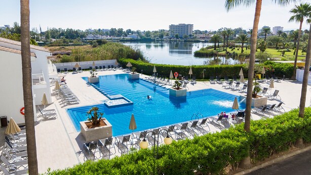 Una piscina con sillones de descanso y sombrillas, rodeada de palmeras y vistas a un lago y edificios.