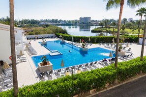 A swimming pool with lounge chairs and umbrellas, surrounded by palm trees and a view of a lake and buildings. - Eix Lagotel Holiday Resort (Muro)