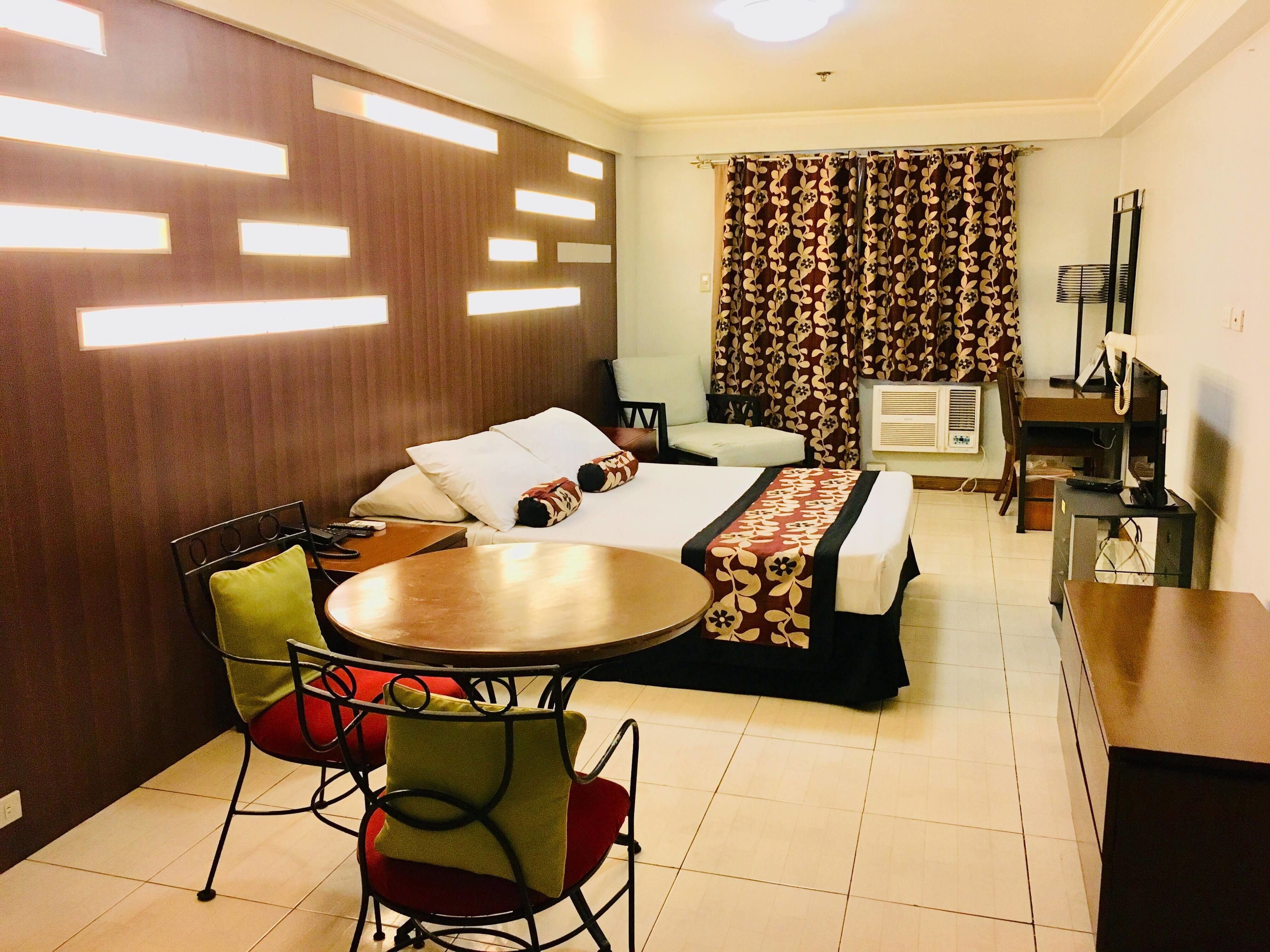 Quarto de hotel com cama, escrivaninha, cadeira, mesa, ar condicionado e janela com cortinas.